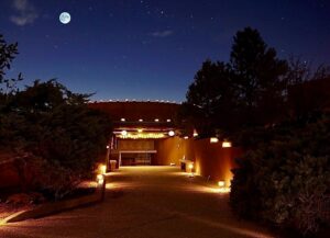 Night view of The Chi Center's Kiva illuminated by soft pathway lights, with dense trees surrounding the area under a star-studded sky and a luminous full moon.