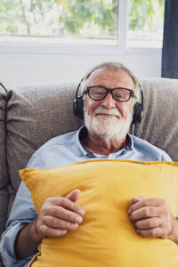 Elderly Caucasian man with a full white beard and glasses smiles contentedly while reclining on a gray sofa, clutching a bright yellow pillow. He wears a blue shirt and large black headphones, participating in a Wisdom Healing Qigong session in his cozy, sunlight-filled living room.
