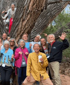 A joyful group of people gathers around a massive, ancient tree, smiling and posing with playful, enthusiastic expressions. Some participants stand near the trunk, while others climb onto the tree’s large branches. The scene radiates warmth, community, and a deep connection to nature, capturing the spirit of gratitude and celebration on a Magical Sunday.