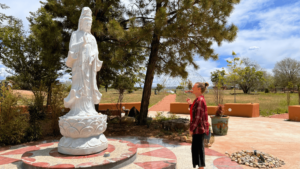A woman stands in quiet contemplation before a large white statue of a serene figure, set in a peaceful outdoor area surrounded by trees. The scene is bathed in soft sunlight, and a path lined with greenery stretches into the background, leading to a distant structure. This moment captures a sense of reverence, mindfulness, and spiritual connection, reflecting the gratitude and tranquility of a Magical Sunday.