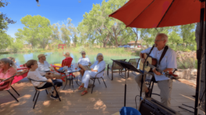 A musician serenades guests on Magical Sunday by a peaceful pond under the shade of a red umbrella. Participants relax in chairs, enjoying the music and the serene natural surroundings, with trees and a reflective pond creating a tranquil atmosphere.