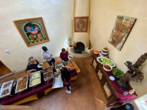 An overhead view of a buffet-style meal setup at Magical Sunday, with participants serving themselves from a variety of dishes, including fresh vegetables, salads, and warm entrees. The setting is a warmly decorated room with spiritual artwork and statues, creating a welcoming atmosphere.