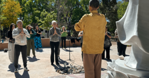 A group of people participate in a guided meditation or Qigong practice outdoors, led by Master Mingtong Gu. The participants stand in a circle with hands in a prayer or meditative position, surrounded by lush greenery and dappled sunlight. A statue and a circular arrangement of stones enhance the serene, sacred atmosphere, capturing the spirit of peace, mindfulness, and inner transformation central to the 2024 Fall Retreats.
