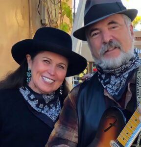 Members of Roadside Distraction, a duo dressed in Western-inspired attire with black hats, turquoise earrings, and patterned bandanas, smiling warmly. One holds a guitar, adding a musical touch to the friendly and inviting atmosphere.
