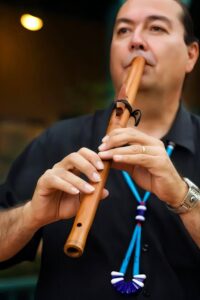 Ronald Roybal playing a wooden Native American flute, holding it gracefully with both hands, focusing on the music. His eyes appear soft, perhaps closed, as he connects deeply with the sound, emanating a peaceful and introspective aura. The warm background lighting enhances the intimate atmosphere, reflecting his soulful connection with the music and his cultural heritage.