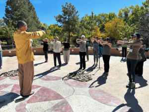 Group practicing Qigong outdoors during the 2024 Fall Retreat, led by an instructor in a yellow robe. Participants stand with arms extended, surrounded by natural scenery and autumn trees, under a bright blue sky.