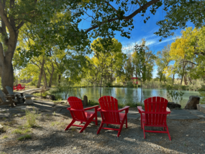 A peaceful lakeside scene with vibrant red Adirondack chairs arranged to face a calm, reflective pond surrounded by lush, green trees under a bright blue sky. Sunlight filters through the branches, casting a warm, inviting glow over the area. The tranquil setting evokes relaxation and contemplation, making it an ideal space for a fall retreat.