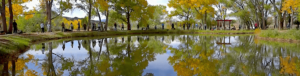 A group of people stand peacefully along the edge of a serene pond, surrounded by trees adorned with vibrant autumn foliage. Their reflections ripple gently on the water, enhancing the tranquil atmosphere. The bright colors of fall and the natural beauty of the setting create a reflective, grounding environment, ideal for a 2024 fall retreat focused on renewal and connection.