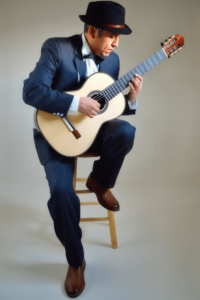 Tito Rios, dressed in a dark suit, white shirt, and black bow tie, sits on a stool playing a classical guitar with focused intensity. He wears a black fedora hat, his gaze directed at the guitar strings as he skillfully performs.