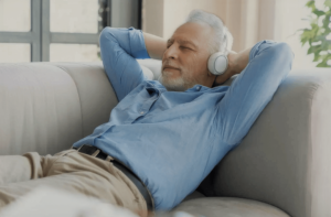 An older man reclining on a sofa with headphones on, eyes closed, and a content expression, lost in a calming audio experience. Dressed in a light blue shirt, he appears relaxed and at peace, symbolizing the rejuvenating and soothing experience offered in the Spring 2024 Retreats - Kiva Healing Livestreams.