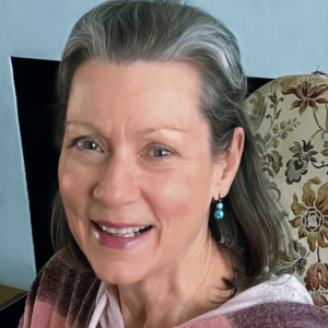 Portrait of Annie, a woman with a warm smile, wearing turquoise earrings and a soft, patterned top. Her gentle expression and presence convey kindness and wisdom, with an elegant touch of silver in her hair, set against a cozy, floral background.