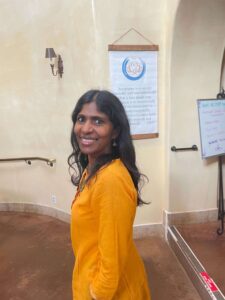 Anusha smiling warmly, wearing a vibrant yellow top, standing indoors against a softly lit background with inspirational decor, radiating positivity and openness.
