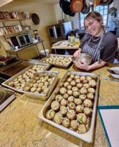 Cheryl Casden Carpenter smiling in a cozy kitchen, surrounded by trays of freshly prepared stuffed mushrooms, showcasing her culinary skills and joy in preparing delicious meals.