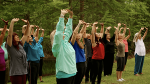 Group of people practicing Qigong outdoors in a lush, green setting, standing with arms raised toward the sky, embodying unity, energy flow, and connection with nature as part of a journey to health and spiritual growth.
