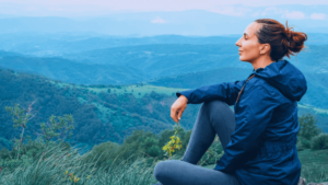 A woman sits peacefully on a grassy hillside, gazing into the distance over expansive green valleys and rolling mountains. Dressed in a blue jacket, she holds a small bundle of wildflowers, embodying a serene connection to nature, self-reflection, and mindfulness.