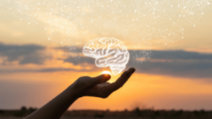 An open hand gently holds an illuminated brain illustration against a sunset sky, symbolizing the power of the mind and the interconnectedness of thought, wisdom, and consciousness. Light connections extend from the brain, representing neural pathways and the limitless potential for learning and growth in the journey of healing and awakening.