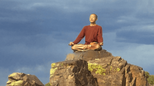 Master Mingtong Gu sits peacefully on a rocky peak under a vast blue sky, eyes closed and hands resting on his knees in a meditative posture. His calm expression and grounded presence reflect a deep connection to nature, embodying serenity and inner harmony—an invitation to embrace stillness and spiritual awakening."
