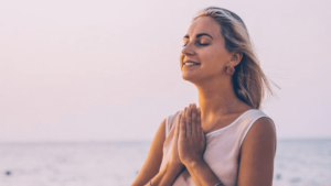 Young woman standing by the sea with hands in prayer position, eyes closed, and a peaceful smile, basking in soft natural light. The image exudes tranquility, gratitude, and mindfulness, symbolizing the inner calm and spiritual connection fostered in the Masterclass
