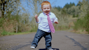 Toddler with a joyful expression walking confidently down a sunlit path, wearing a white shirt, jeans, and a playful pink tie. The image conveys innocence, enthusiasm, and a carefree spirit, symbolizing fresh energy and new beginnings.