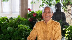 A warm portrait of Master Mingtong Gu, dressed in a beautifully detailed golden traditional garment, exuding a sense of peace and joy. His gentle smile and clear, compassionate gaze convey kindness and wisdom. He is surrounded by lush green plants and vibrant red flowers, with a serene Buddha statue in the background, adding a spiritual ambiance to the setting. This image captures Master Mingtong Gu’s connection to nature, mindfulness, and his role as a guide in healing and spiritual growth.