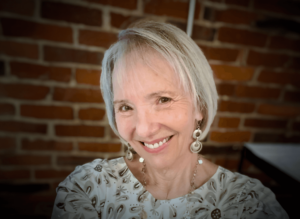 Close-up of Sallena Pool smiling warmly in front of a brick wall backdrop. She has short, light gray hair and is wearing decorative earrings and a patterned top, exuding a joyful and friendly presence.