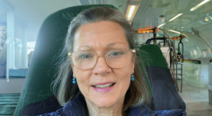 A close-up of Annie, smiling warmly while seated in a modern train or transit setting. Wearing glasses and blue earrings, she exudes a calm and approachable presence, with soft lighting creating a serene, candid moment.
