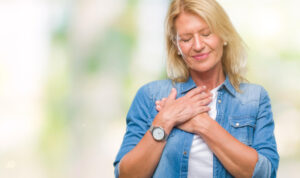 Middle-aged woman with closed eyes and a peaceful expression, gently holding her hands over her heart. Dressed in a denim shirt with a soft, natural light background, she embodies a moment of gratitude and inner connection, symbolizing the heart-centered focus of Qigong practice and the union of health and spiritual growth