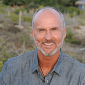 Chip Conley smiles warmly in an outdoor setting, wearing a casual grey shirt. His expression is friendly and approachable, with a neatly trimmed beard and a shaved head. The natural, earthy background suggests a calm and grounded presence, reflecting his work in fostering wisdom and community.