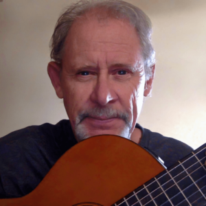 A portrait of Chris Harrell, a man with a gentle, thoughtful expression, holding a classical guitar close to his face. His silver hair and neatly trimmed mustache add to his distinguished appearance, while his gaze conveys warmth and introspection. The soft, neutral background highlights his connection with the instrument, suggesting a deep passion for music and artistic expression.