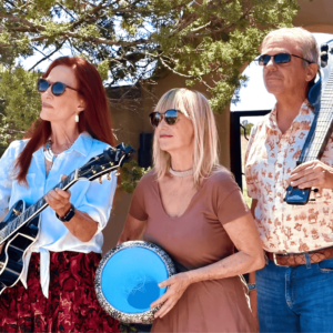 A photo of the Corn Music Band trio outdoors under a sunny sky. The three band members, wearing sunglasses and casual attire, hold their instruments—an electric guitar, a frame drum, and a unique stringed instrument. They stand in a serene, natural setting with green trees in the background, exuding a relaxed, harmonious vibe as they prepare to share their music.
