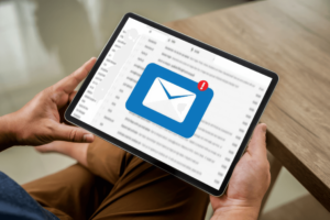 A person holding a tablet displaying an email inbox, with a large envelope icon and a red notification badge indicating a new message. The email icon is prominently centered on the screen, symbolizing communication and connectivity. The background shows a blurred list of emails, highlighting the focus on a new, unread message. This image conveys the theme of digital communication and staying connected through email notifications.