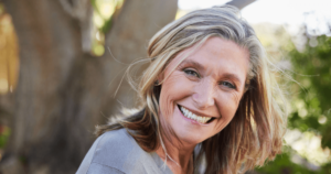 Middle-aged woman with a warm, joyful smile, outdoors in natural sunlight, surrounded by greenery, embodying health, vitality, and contentment, symbolizing the benefits of Wisdom Healing Qigong for health and healing.