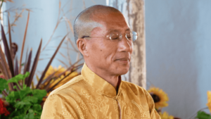 Master Mingtong Gu meditating peacefully with closed eyes, wearing a golden traditional garment adorned with intricate patterns, set against a soft, natural background with greenery and flowers.