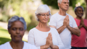 Group of diverse adults meditating outdoors, with eyes closed and hands in prayer position, led by an elderly woman in the center with silver hair and a peaceful expression, embodying mindfulness, unity, and spiritual growth.
