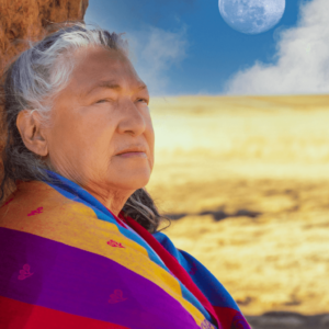 Grandmother Flordemayo stands outdoors against a natural rock, gazing thoughtfully into the distance. She is wrapped in a vibrant shawl with rich colors of red, purple, and blue. The landscape around her is expansive and sunlit, with a bright blue sky and a softly glowing moon above. Her serene expression and the earthy, sacred setting convey a deep connection to nature and wisdom