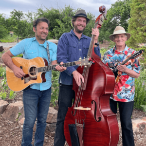 The High Desert Trio performing outdoors, with each musician holding an instrument: one with an acoustic guitar, one with a double bass, and the third with a mandolin. All three are smiling, dressed in casual, colorful attire, and surrounded by natural greenery. The image captures a lighthearted and joyful moment, reflecting the trio's camaraderie and passion for music.