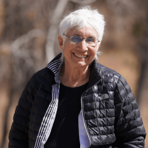 Judy Tuwaletstiwa smiles warmly while outdoors, wearing glasses, a black North Face jacket, and a striped collar shirt. Her white hair and relaxed expression convey a sense of wisdom and kindness. The natural, softly blurred background adds to the serene and grounded feeling of the image, reflecting her artistic and contemplative nature.