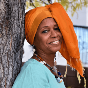 Karen Jones Meadows leans against a tree with a warm, friendly smile. She wears a vibrant orange head wrap and a light turquoise blouse, complemented by a unique, multi-beaded necklace. Her expression exudes calm and joy, embodying a spirit of wisdom and resilience. The natural setting and her colorful attire highlight her connection to cultural roots and personal strength.