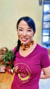 A portrait of Linling, smiling warmly and standing indoors. She wears a purple T-shirt with the gold logo of the Chi Center and a multi-layered beaded necklace in earthy tones. Her hair is styled in a side braid, and she stands against a softly blurred background, exuding a sense of warmth and positivity.