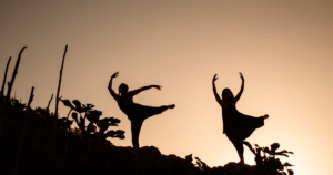 Two silhouetted figures dance gracefully atop a hill at sunset, their arms and legs extended in expressive poses against a warm, glowing sky. Surrounding plants add to the natural beauty of the scene. This image captures a sense of freedom, connection to nature, and the transformative energy of collective awakening, embodying the spirit of Living Tao.