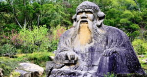 A majestic stone statue of a wise, ancient sage seated amidst lush greenery and vibrant blossoms, embodying the essence of deep wisdom and timeless presence. The sage's calm, contemplative expression and flowing beard evoke reverence and spiritual depth. Surrounded by trees and flowering plants, this setting harmonizes nature with the eternal, symbolizing the journey of Living Tao: A Journey of Collective Awakening and Profound Transformation. This image captures the peaceful, grounded energy of Taoist philosophy, inviting viewers into a space of reflection, insight, and connection with the natural world.