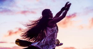 A powerful image of a woman silhouetted against a pastel-colored sunset sky, her arms raised gracefully upward in a gesture of reverence or celebration. Her long, flowing hair and traditional, patterned attire capture the essence of freedom, connection, and cultural reverence. This evocative scene, set against the warm and serene hues of dusk, embodies the spirit of Living Tao: A Journey of Collective Awakening and Profound Transformation, symbolizing unity with nature, spiritual awakening, and the deep, transformative power of mindful movement and tradition.
