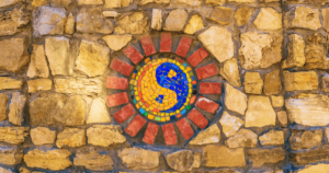 A vibrant mosaic embedded in a rustic stone wall, featuring a dynamic yin-yang symbol in blue and yellow. Surrounding the yin-yang are colorful tiles in shades of red, orange, green, and yellow, arranged in a circular pattern, evoking a sense of balance, harmony, and radiant energy. The stone wall adds an earthy, grounded quality, symbolizing ancient wisdom and timelessness. This image beautifully represents Living Tao: A Journey of Collective Awakening and Profound Transformation, capturing the essence of unity, interconnectedness, and the fusion of spiritual insight with grounded practice.