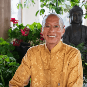 A warm portrait of Master Mingtong Gu, dressed in a beautifully detailed golden traditional garment, exuding a sense of peace and joy. His gentle smile and clear, compassionate gaze convey kindness and wisdom. He is surrounded by lush green plants and vibrant red flowers, with a serene Buddha statue in the background, adding a spiritual ambiance to the setting. This image captures Master Mingtong Gu’s connection to nature, mindfulness, and his role as a guide in healing and spiritual growth.