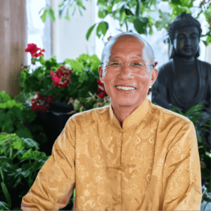 A warm portrait of Master Mingtong Gu, dressed in a beautifully detailed golden traditional garment, exuding a sense of peace and joy. His gentle smile and clear, compassionate gaze convey kindness and wisdom. He is surrounded by lush green plants and vibrant red flowers, with a serene Buddha statue in the background, adding a spiritual ambiance to the setting. This image captures Master Mingtong Gu’s connection to nature, mindfulness, and his role as a guide in healing and spiritual growth.