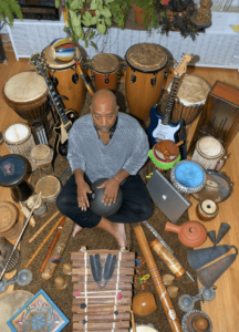 Maurice, a master musician, sits cross-legged amidst a rich collection of musical instruments from around the world. He is centered, holding a drum, surrounded by congas, guitars, flutes, drums, and various percussion instruments, creating a vibrant scene of musical diversity. The setting, decorated with lush plants, reflects a harmonious blend of culture, rhythm, and artistry.