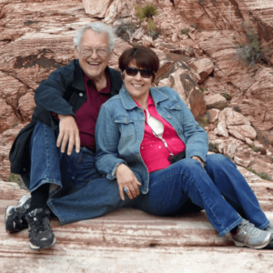 Rick and Judy sitting together on rugged, red rock terrain, sharing a joyful moment. Rick, with white hair and glasses, is wearing a dark jacket over a red shirt, while Judy, with short dark hair and sunglasses, sports a denim jacket over a bright pink shirt. Both are smiling warmly, exuding a sense of companionship and happiness amidst the natural landscape, capturing the spirit of connection and community often felt in a drum circle gathering.