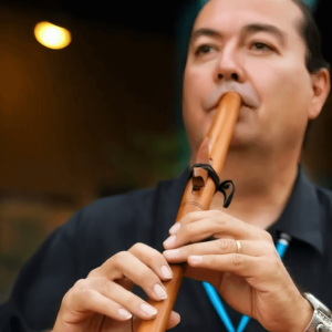 Ronald Roybal playing a wooden Native American flute, holding it gracefully with both hands, focusing on the music. His eyes appear soft, perhaps closed, as he connects deeply with the sound, emanating a peaceful and introspective aura. The warm background lighting enhances the intimate atmosphere, reflecting his soulful connection with the music and his cultural heritage.