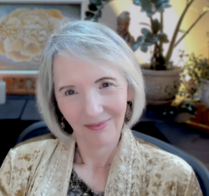 Portrait of Sallena Pool, smiling warmly in a peaceful indoor setting with soft lighting, wearing a patterned golden shawl, symbolizing compassion, wisdom, and gentle guidance as part of the Passages coaching program.