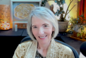 Portrait of Sallena Pool, smiling warmly in a peaceful indoor setting with soft lighting, wearing a patterned golden shawl, symbolizing compassion, wisdom, and gentle guidance as part of the Passages coaching program.